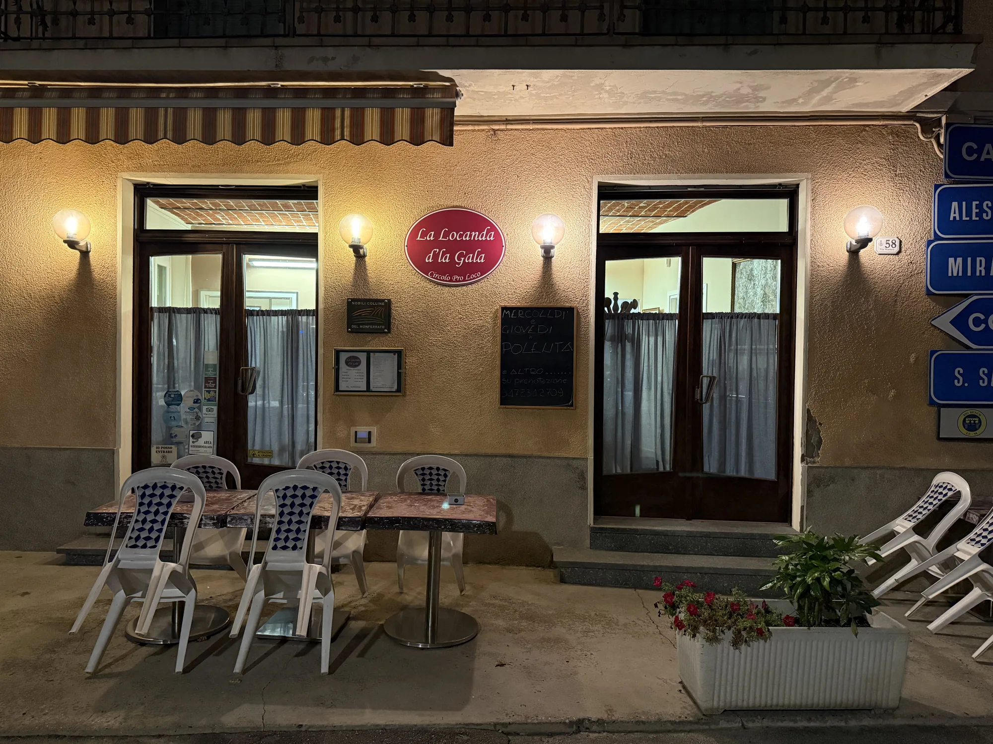 L'ingresso della Locanda d'la Gala di sera: l'insegna ovale bordeaux tra i lampioni accesi, la lavagna con le serate del mercoledì e del giovedì e i tavolini sulla strada.